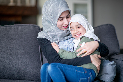 A beautiful Muslim mother spending time with her four year old daughter at home. They are both wearing a hijab. The mother is snuggling her daughter who is sitting on her lap.