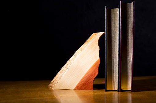 Sleek artistic sculpture made of Cedar sits on a golden oak table top.   Black background.  Sculpture art used as a book end against leather bound books.