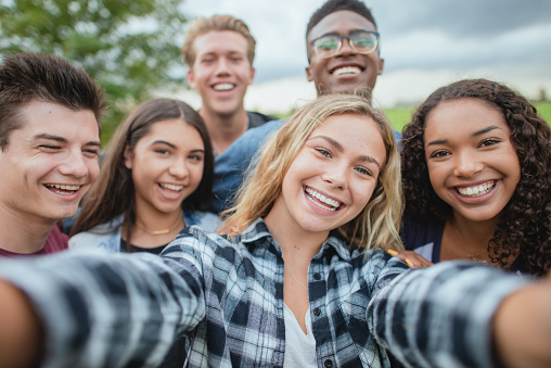 A multi ethnic group of teenagers huddled together for a selfie while standing outside. They are dressed casually. They are happy and smiling.