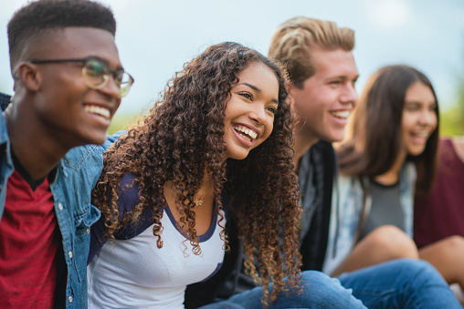 A multi ethnic group of teenagers hanging outdoors together. The focus of the photo is on an African American teenage girl who is smiling and happy to be with her friends.