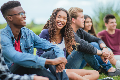 A multi ethnic group of teenagers hanging outdoors together. The focus of the photo is on an African American teenage girl who is smiling and happy to be with her friends.