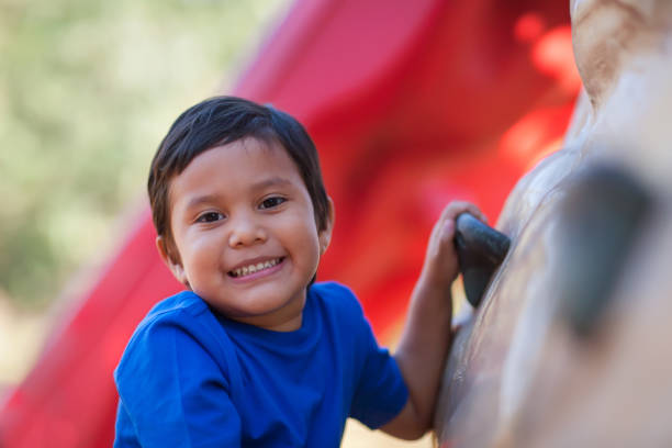 4 year old boy smiles while holding on to a kids outdoor rock climbing wall. stock photo