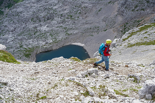 Side view of Mature woman hiking  above 2000 m at one of  the most photogenic locations in Julian Alps.  She is on top of rock, above clouds with helmet on her head, camera in her hands and there is Upper Krisko Lake in background.