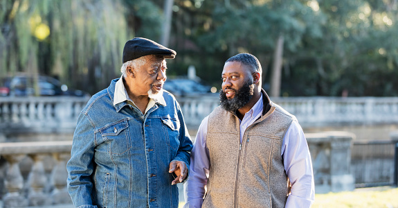 A senior African-American man in his 70s taking a walk with his son-in-law, a mid adult man in his 30s, on a sunny autumn day at a park.