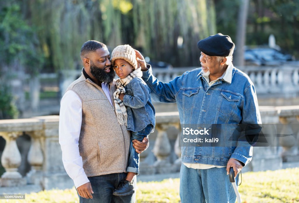 Three generation African-American family walking in park Senior man with his son and grandson taking a walk in the park on a sunny autumn day. The little boy is 3 years old. He is in his father's arms and his 70 year old grandfather is touching the knit hat on his head. Son Stock Photo