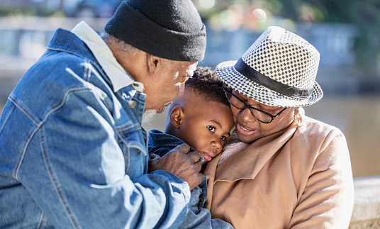 A 3 year old African-American boy in the park with his grandparents on a sunny autumn day. His grandmother, in her 60s, is holding him in her arms. His grandfather, a senior man in his 70s, is looking at him, with his hand on the boy's shoulder.