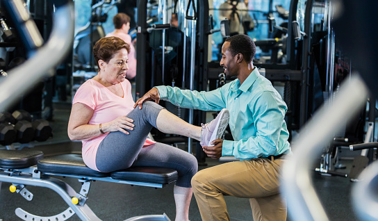 A physical therapist examines a senior woman with a leg injury. They will do exercises in a gym to help her recovery. He is kneeling in front of her, holding her heel as she lifts her leg and bends her knee. The medical professional is African-American and the patient is Caucasian.
