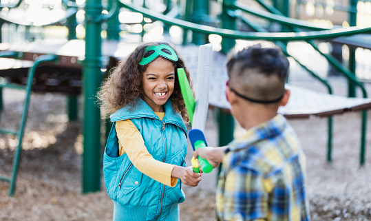 Two multiracial children playing together on the playground, fighting with toy swords. They are siblings, a 7 year old sister and 8 year old brother, mixed race African-American and Caucasian. The view is from over the boy's shoulder and the focus is on the girl.