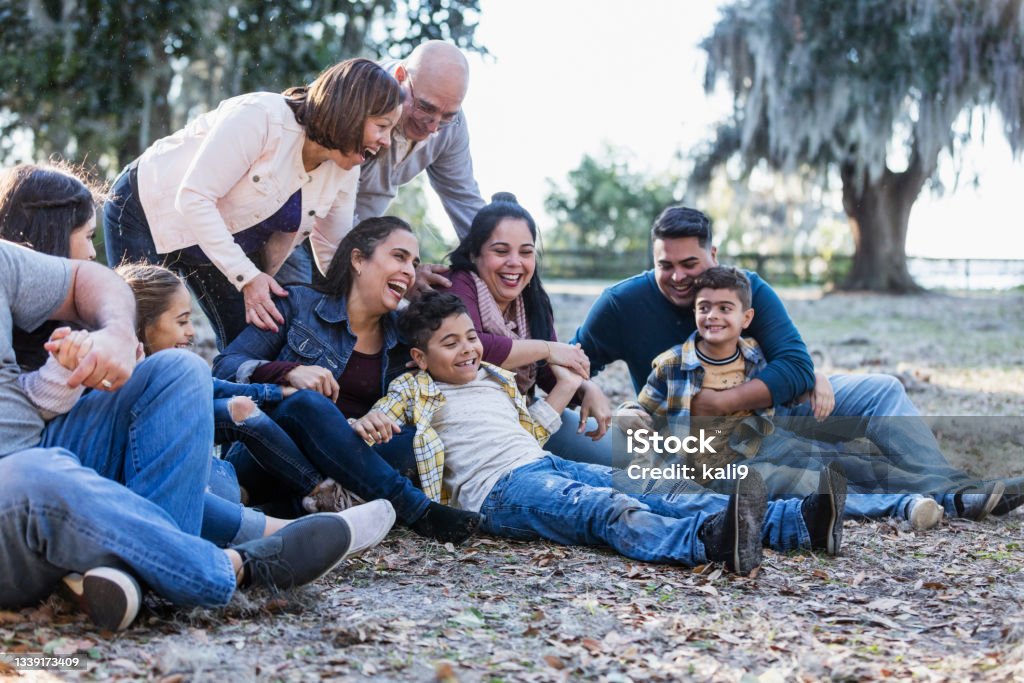 Multi-generation Hispanic family at park, sit on ground A large multi-generation Hispanic family having fun at the park laughing as they sit down together on the ground. The grandparents are still standing, behind their children and grandchildren. The ten people range in age from 7 to early 70s. Multi-Generation Family Stock Photo