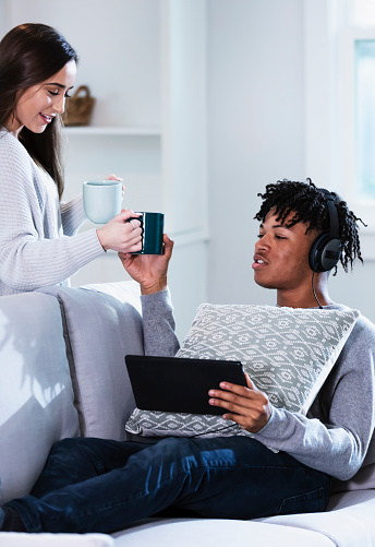 A young multiracial couple relaxing at home together. The African-American man is sitting on the sofa, wearing headphones and using a digital tablet. She is standing behind the sofa smiling and handing him a coffee cup. She is mixed race Hispanic and Caucasian. They are both 19 years old.