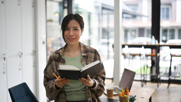 istock Young woman holding book and smiling to camera while standing in modern workplace. 1339188579