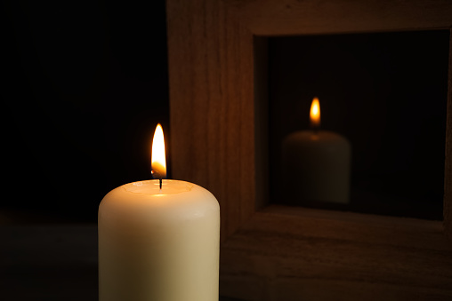 Large white candle burning in the darkness with reflection on glass in a wooden frame in a conceptual image