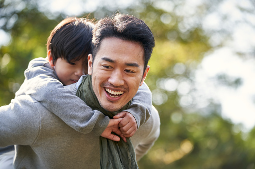 asian father carrying son on back having fun outdoors in park