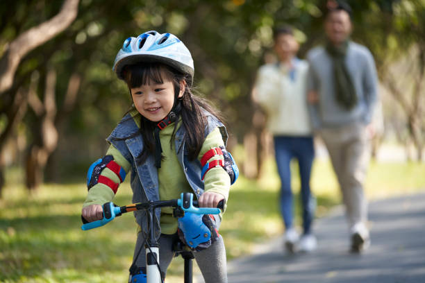 little asian girl riding bike outdoors in park parents watching from behind - helmet bicycle little girls child imagens e fotografias de stock