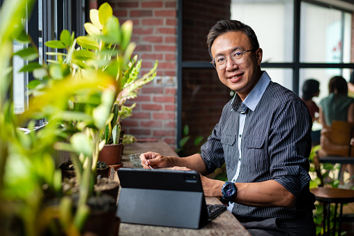 Mature Asian entrepreneur businessman having take away food during lunch break at eco-friendly cafeteria with beautiful potted plants decoration. The businessman looking at camera with digital tablet on table and sitting next to balcony window.