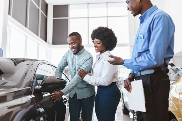 autoverkaufsgeschäft. manager im gespräch mit afro-paar, zeigt ihnen neues auto im autohaus - kaufen stock-fotos und bilder
