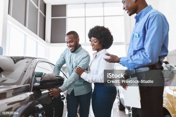Car Sales Business Manager Talking To Afro Couple Showing Them New Auto At Dealership Shop Stock Photo - Download Image Now