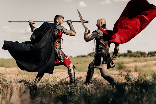 Battle with spear and sword between two ancient greek or roman warriors in battle dress and cloaks on meadow.