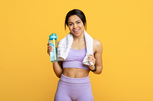 Stay hydrated during workout. Happy african american sportswoman with towel and bottle of water on yellow studio background. Positive fit lady looking at camera and smiling