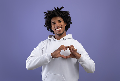 Kind black teenager shaping hands like heart near his chest, making love gesture on violet studio background. Friendly African American teen guy expressing kindness, showing romantic feelings