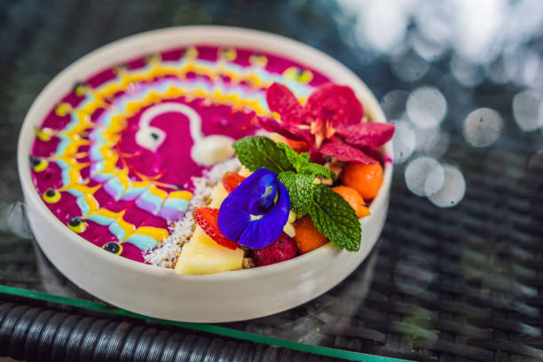 Healthy tropical breakfast, smoothie bowl with tropical fruits, decorated with a pattern of colorful yogurt with turmeric and spirulina. It is also decorated with fruits, flowers, chia seeds, coconut, granola, pineapple, mint and strawberries