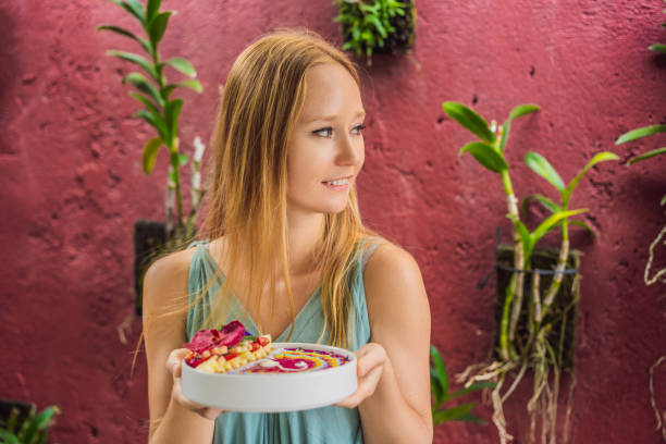 istock Young woman having a mediterranean breakfast, eats Healthy tropical breakfast, smoothie bowl with tropical fruits, decorated with a pattern of colorful yogurt with turmeric and spirulina. It is also decorated with fruits, flowers, chia seeds, coconut 1339221647