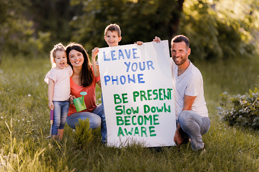 Happy family spending a spring day in nature while father and son are holding a placard with text 'Leave your phone, be present, slow down and become aware'.