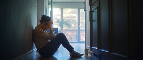 joven triste sentada en el suelo en el pasillo de su apartamento, cubriéndose la cara con las manos. atmósfera de depresión, problemas en la relación, muerte en la familia. dramático momento de malas noticias - víctima fotografías e imágenes de stock