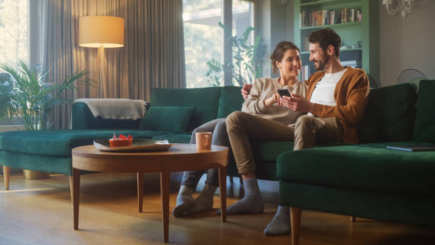 カップルは、居心地の良いアパートのソファに座っている間、スマートフォンデバイスを使用しています。ボーイフレンドとガールフレンドの話は、インターネット上で電子ショッピングを� - 住宅 ストックフォトと画像