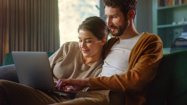 istock Young Couple Use Laptop Computer, while Sitting on a Couch in the Cozy Stylish Apartment. Boyfriend and Girlfriend Shopping on Internet, Using Social Media, Watching Funny Videos and Streaming Service 1339226445