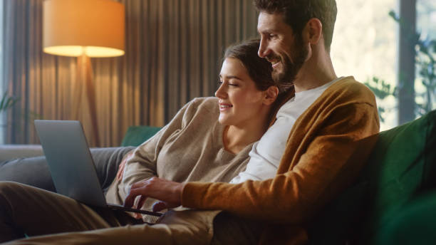 istock Young Couple Use Laptop Computer, while Sitting on a Couch in the Cozy Stylish Apartment. Boyfriend and Girlfriend Shopping on Internet, Using Social Media, Watching Funny Videos and Streaming Service 1339226465