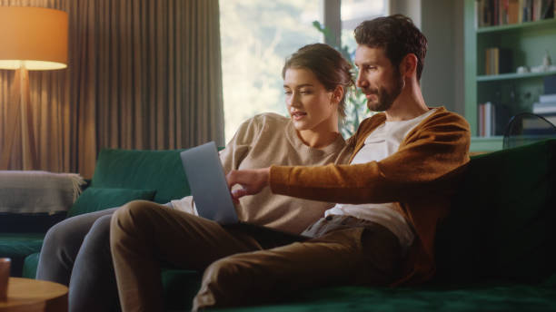 istock Young Couple Use Laptop Computer, while Sitting on a Couch in the Cozy Stylish Apartment. Boyfriend and Girlfriend Shopping on Internet, Watching Videos and Streaming Service. 1339226486