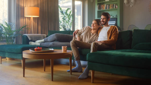 paar schaut zusammen fernsehen, während sie auf einer couch im wohnzimmer sitzen. freundin und freund umarmen, kuscheln, reden, lächeln und schauen tv-streaming-dienste. haus mit gemütlichem stilvollem interieur. - pärchen tv stock-fotos und bilder