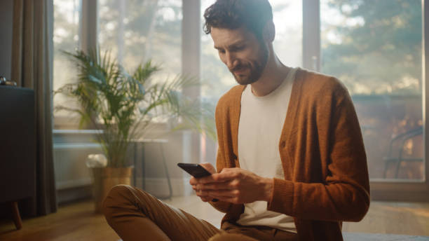 istock Man Using Smartphone at Home, Does Remote Work. Handsome Smiling Male Sitting on the Floor Works with Papers, Documents, Brainstorms Creative Project with Online Research. Sunny Living Room 1339226628