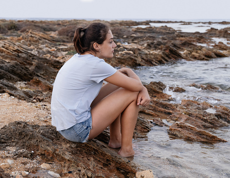 Thoughtful teenage girl sitting on cliff by the sea with legs in water at sunset and looking straight, wearing blue t-shirt and jeans shorts, side view. T-shirt mockup