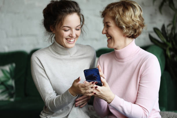 famille et technologie. une femme âgée et sa fille adulte utilisent un smartphone à la maison - grandparent senior adult child reading photos et images de collection