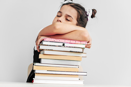 Bored and tired schoolgirl sleeping on a pile of books on the desk against light grey background. Exhausted little girl learner falling asleep during reading lot of books. Consuming education concept.