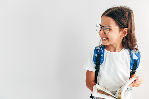 Horizontal image of a little girl in transparent eyeglasses with backpack smiling looking at one side with copy space. School kid with backpack and book has joyful expression to going back to school.