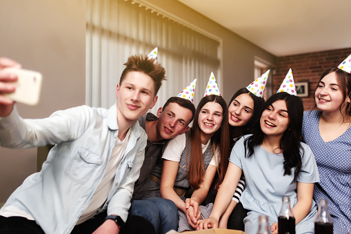 Group of best friends with festive cap taking selfie at birthday party. Girls and guys are posing and smiling at camera of mobile phone. High quality photo