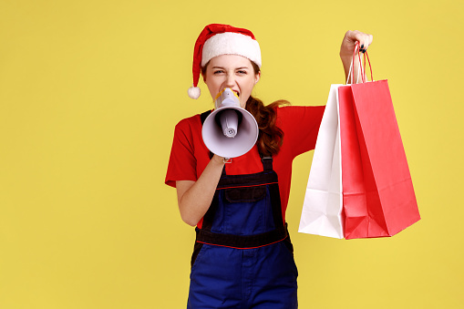 Female courier screams in megaphone, showing shopping bags, announcing discounts of delivery service, wearing blue overalls and santa claus hat. Indoor studio shot isolated on yellow background.