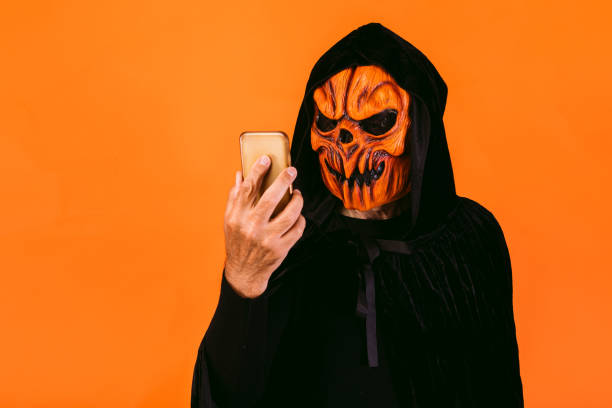 Man dressed in pumpkin latex mask and hooded velvet cape, looks at his mobile phone. Halloween and days of the dead concept. stock photo