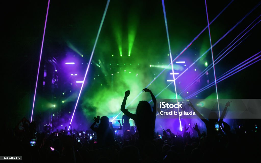Rave party silhouettes. Rear view of large group of people enjoying a concert performance. There are many hands applauding and taping the show. Multi colored lasers and spot lights firing from the stage.
Silhouettes have been significantly liquified. Green Color Stock Photo