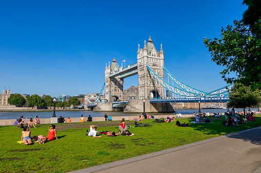 London, United Kingdom - 07 Sep 2021. Londoners and tourists enjoying the summer sunshine sitting on the grass with Tower Bridge stretching across the River Thames in the background against a clear blue sky.