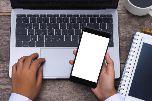 istock Mock up image of business person hand using black mobile smart phone with blank white screen and work on laptop computer with cup of coffee on wooden table at cafe. Top view. Work at home concept. 1339261317