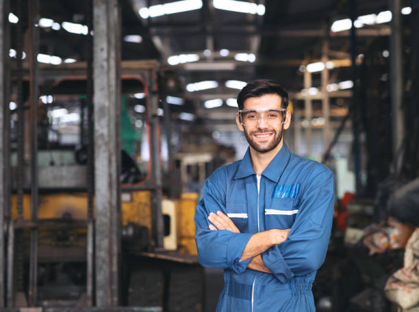 lächelnder arbeiter in schutzbrille stehend mit gekreuztem arm in der nähe von maschinen zur herstellung von industrieanlagen in der fabrik - manager foreman warehouse arms crossed stock-fotos und bilder