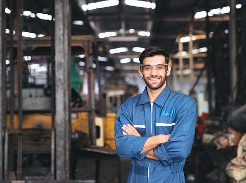 Smiling worker in safety glassess standing with arm crossed near industrial equipment manufacturing machinery in the factory