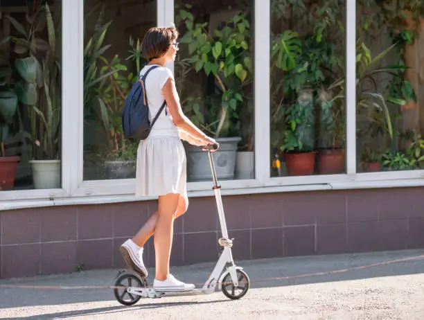Photo of Young woman rides kick scooter down the street at sunset. Tropical plants for landscaping and indoor gardening in window of flower shop. Eco-friendly transport. Healthy lifestyle.