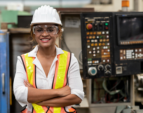 Portrait of confident maintained woman worker standing with arm crossed in front of manufacturing machinery in the factory