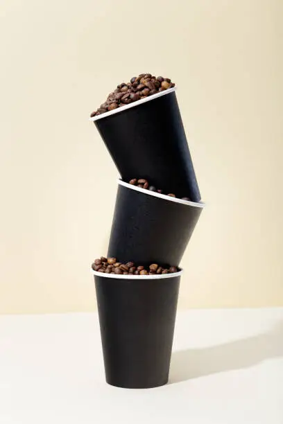 Three black paper coffee cups filled with coffee beans, beige background