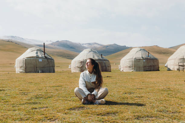 Woman traveler enjoying sunny summer day at the yurt camp in the mountains of Kyrgyzstan Young woman with long hair sitting at the meadow near the nomad yurts with mountain view in Tian Shan, Central Asia national heritage site stock pictures, royalty-free photos & images